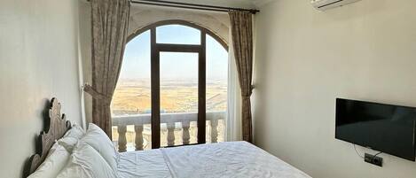 A bedroom with a bed, a television, an air conditioning unit, a chandelier, and a window with a view.