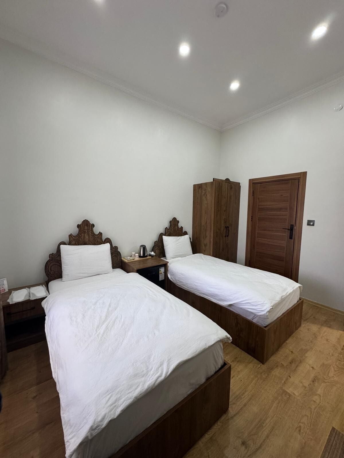 A bedroom with two wooden beds, white bedding, a nightstand with a lamp, and a wooden wardrobe.