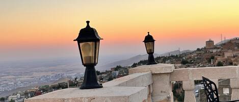 A terrace with two street lamps, a chair, and a view of a cityscape at sunset.