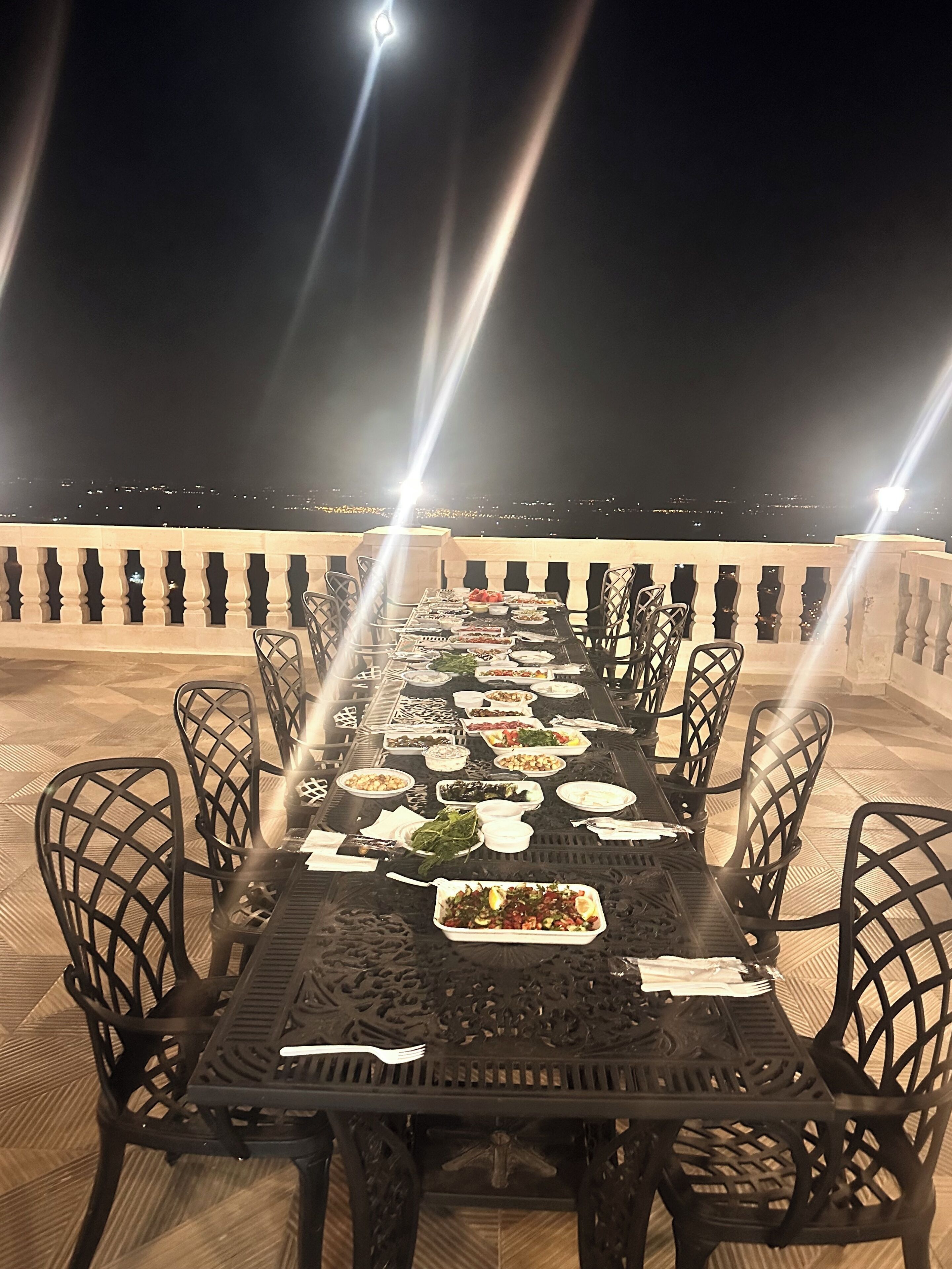 Una larga mesa de comedor lista para una comida en una terraza al aire libre con vista a la ciudad por la noche.