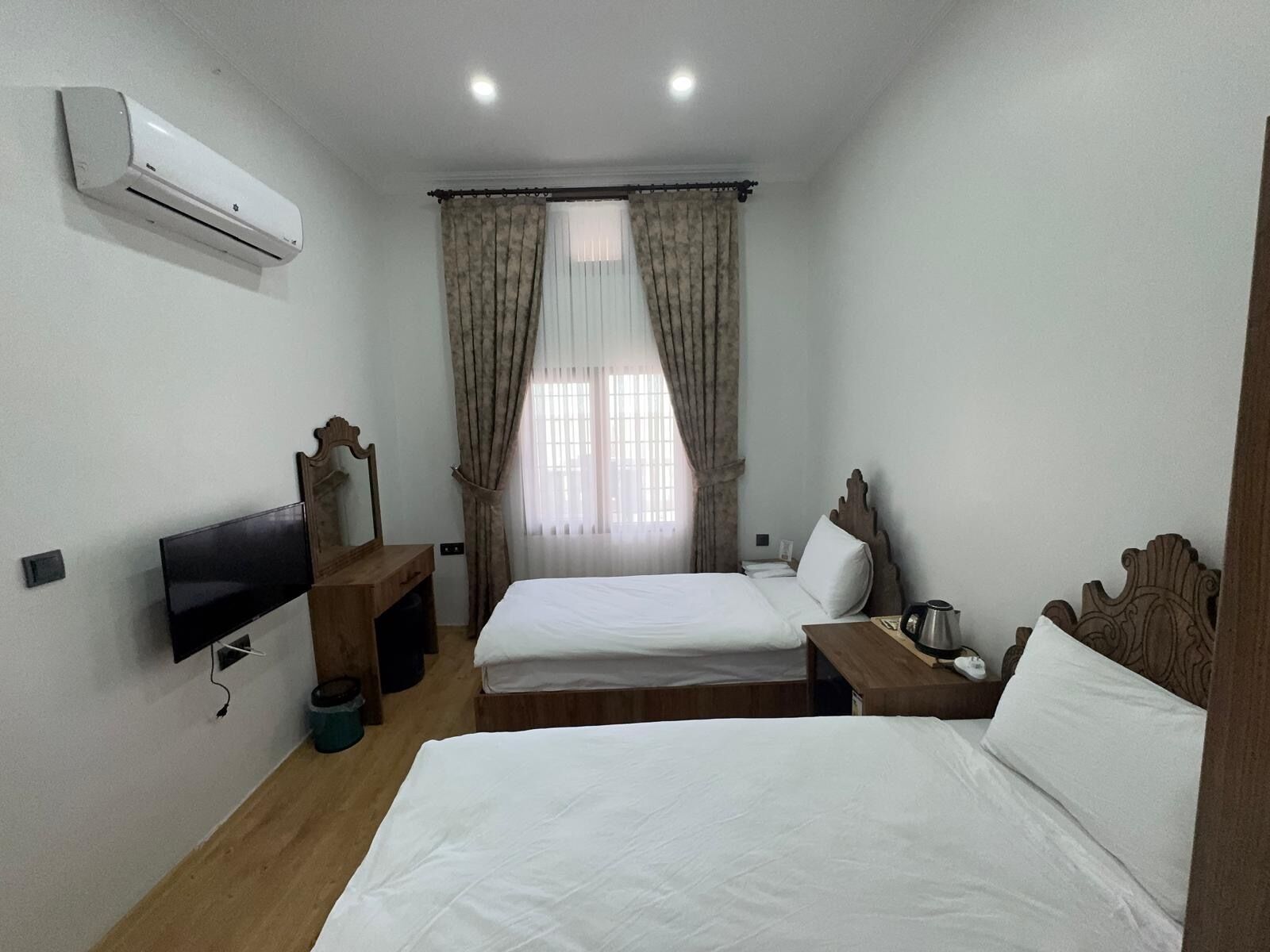 A hotel room with two beds, a TV, a wooden dresser, a window with curtains, and an air conditioning unit.
