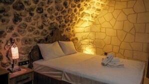 A bed with white linens and a headboard, a bedside lamp, and a stone wall. - BABİ NEFES Demirhan Konağı (Mardin)