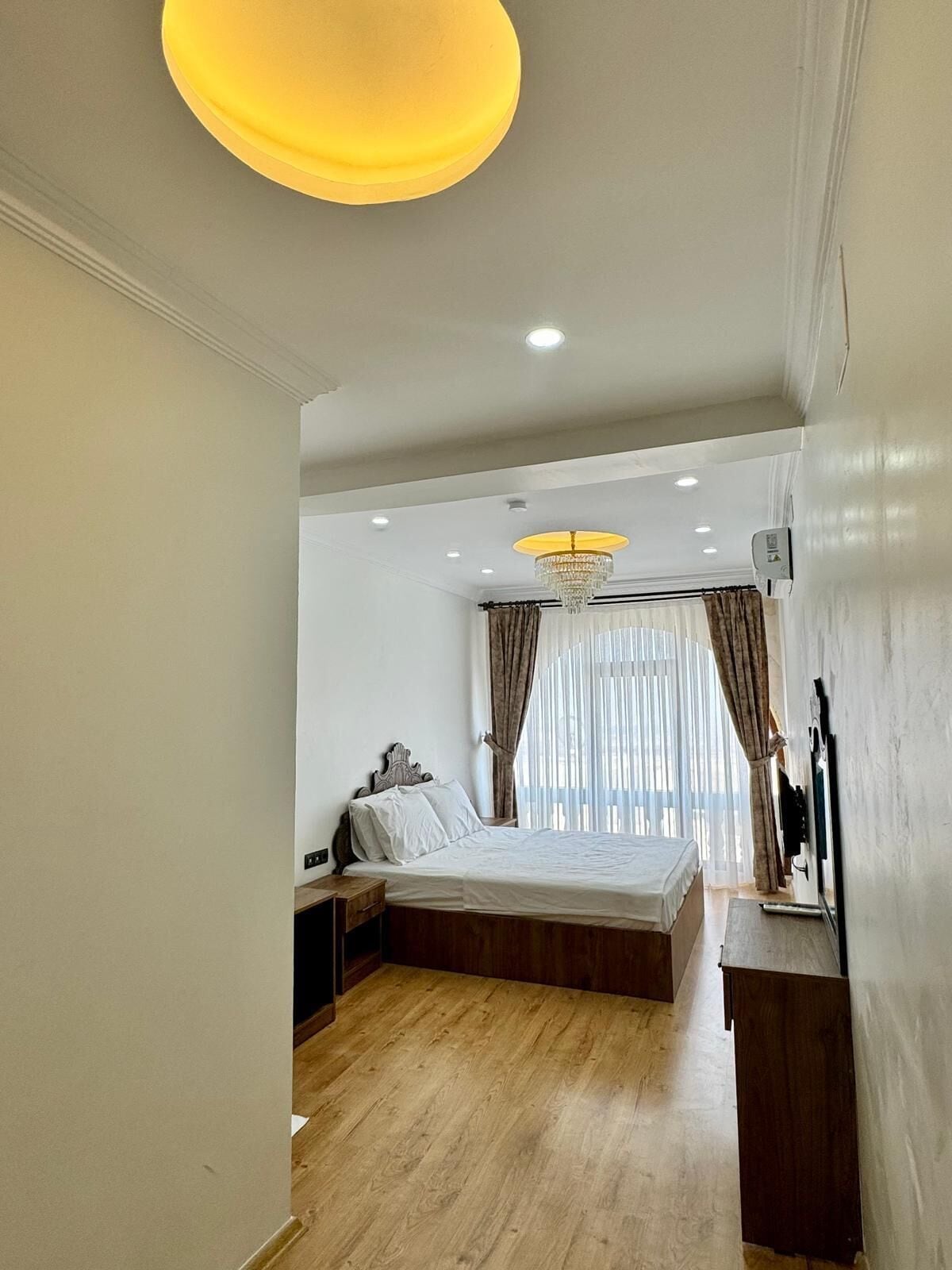 A bedroom with a large bed, wooden flooring, a chandelier, and a window with curtains.