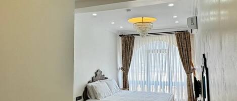 A bedroom with a large bed, wooden flooring, a chandelier, and a window with curtains.