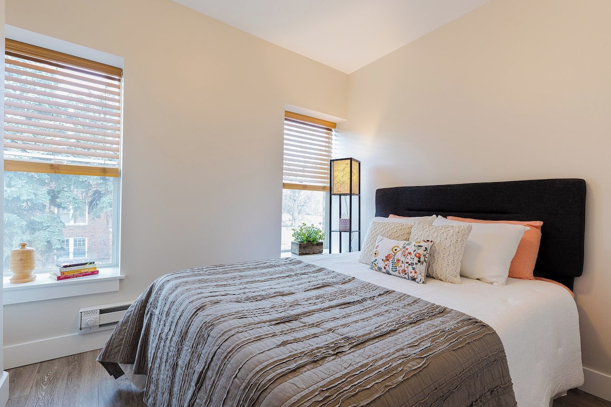 A bedroom with a bed, a nightstand, a window with blinds, and a view of the outside.