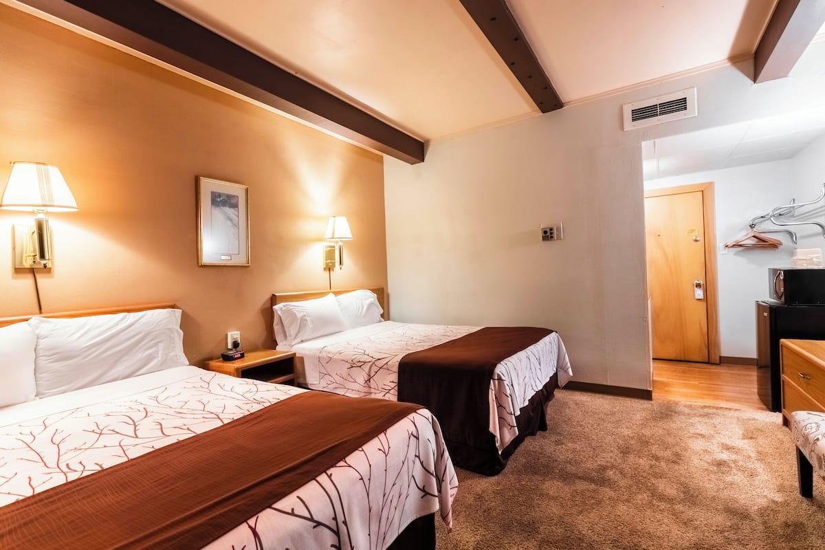 A hotel room with two beds, each with a bedside table and lamp, a wooden headboard, a framed picture on the wall, and a door leading to another room.