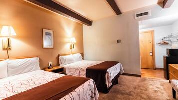 Habitación de hotel con dos camas, cada una con una mesita de noche y lámpara, cabecero de madera, un cuadro enmarcado en la pared y una puerta que da a otra habitación.