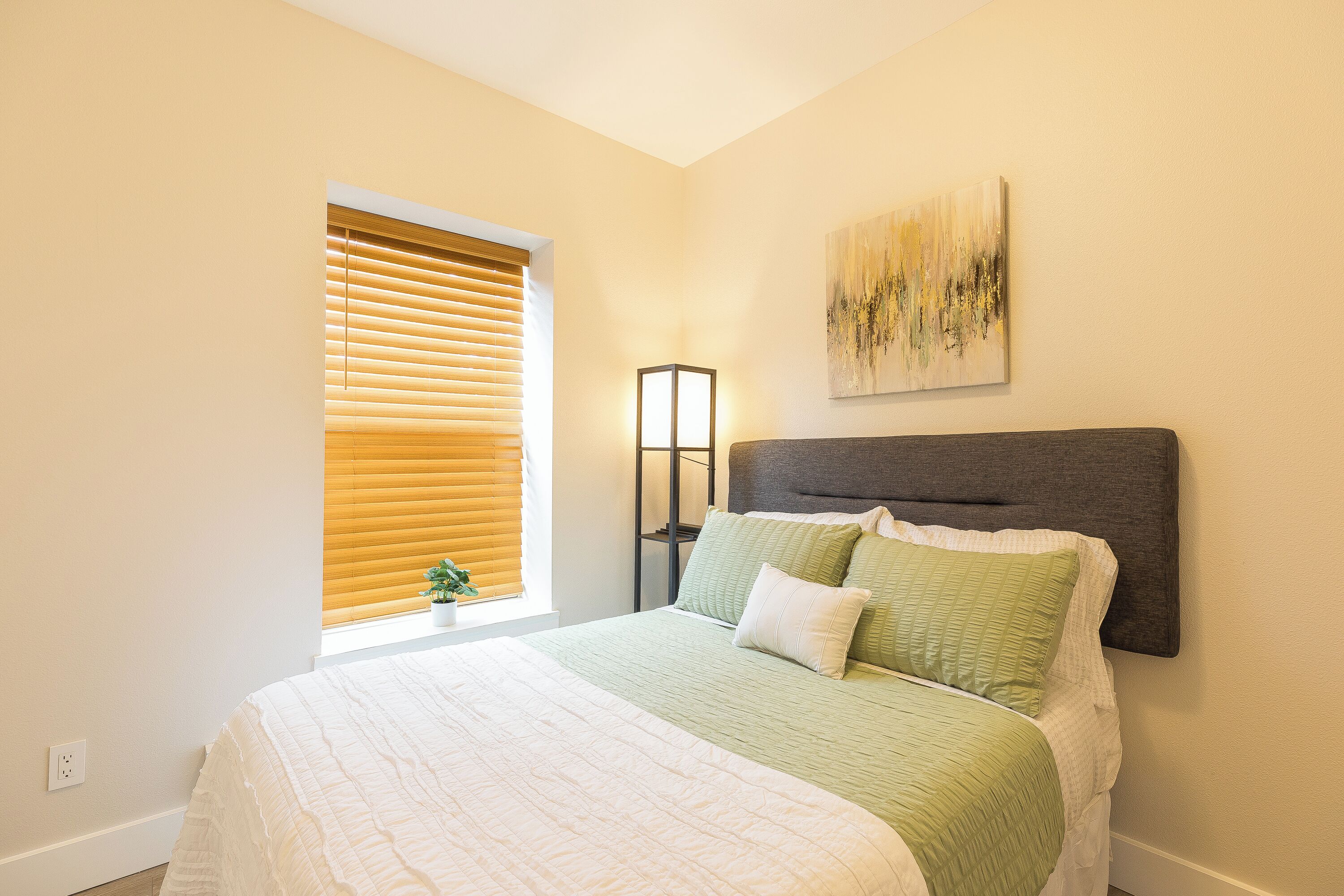 A bedroom with a bed, a bedside table, a lamp, a window with blinds, and a painting on the wall.