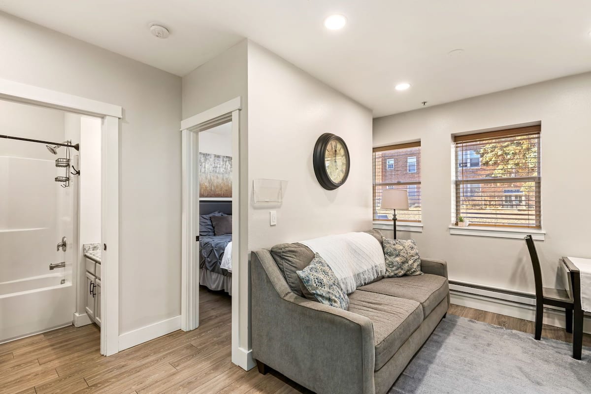 A modern apartment with a gray sofa, a clock on the wall, and a window with blinds.