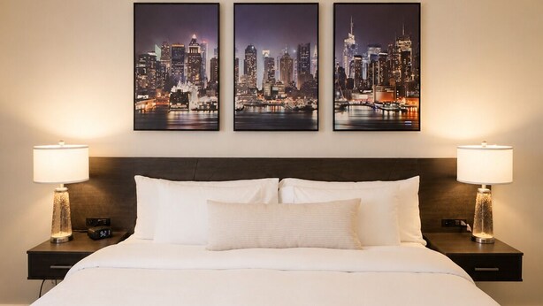 Habitación de hotel con una cama grande, dos mesitas de noche con lámparas y tres cuadros con vistas urbanas en la pared.
