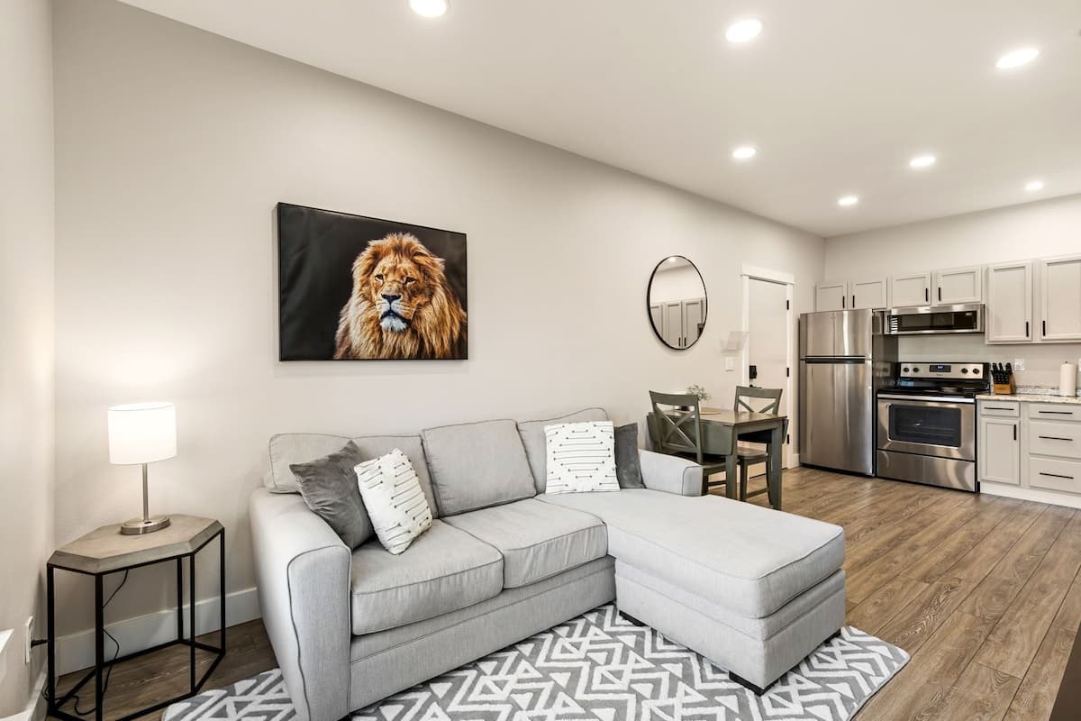 A modern living room with a grey sectional sofa, a dining table, and a kitchen area with stainless steel appliances.