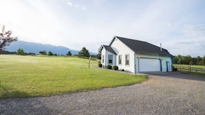 Une maison de plain-pied avec un garage, entourée d’un champ verdoyant et avec des montagnes en arrière-plan.