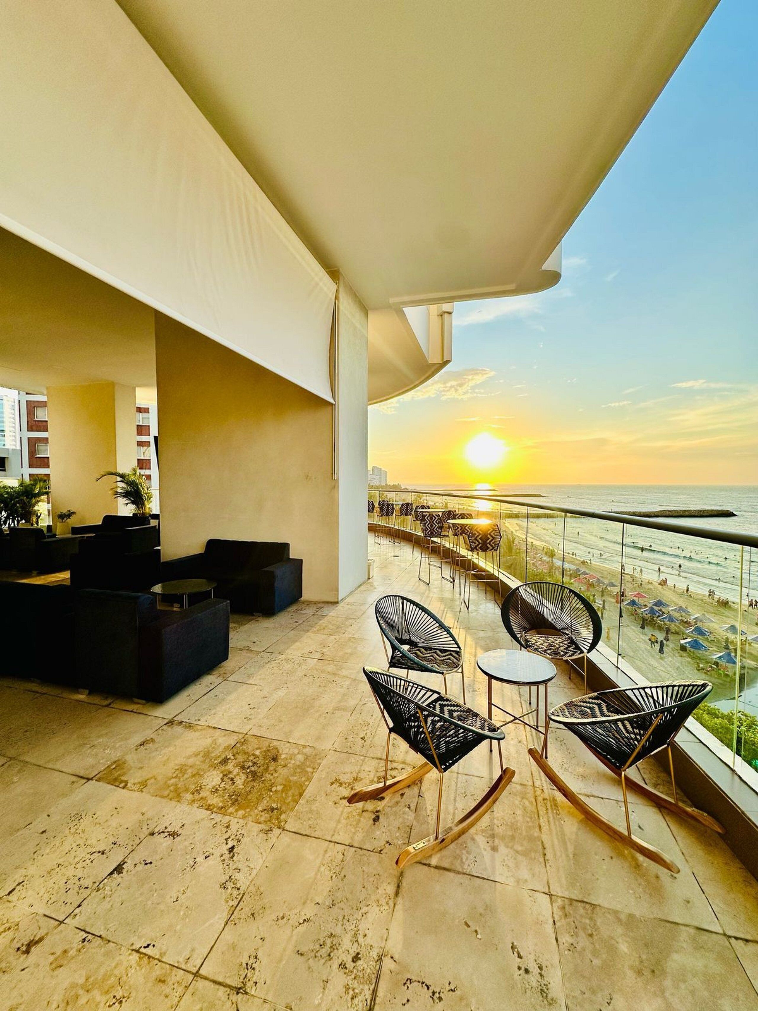 A balcony with outdoor seating, a view of the sunset over the water, and a cityscape in the background.