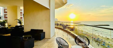 A balcony with outdoor seating, a view of the sunset over the water, and a cityscape in the background.