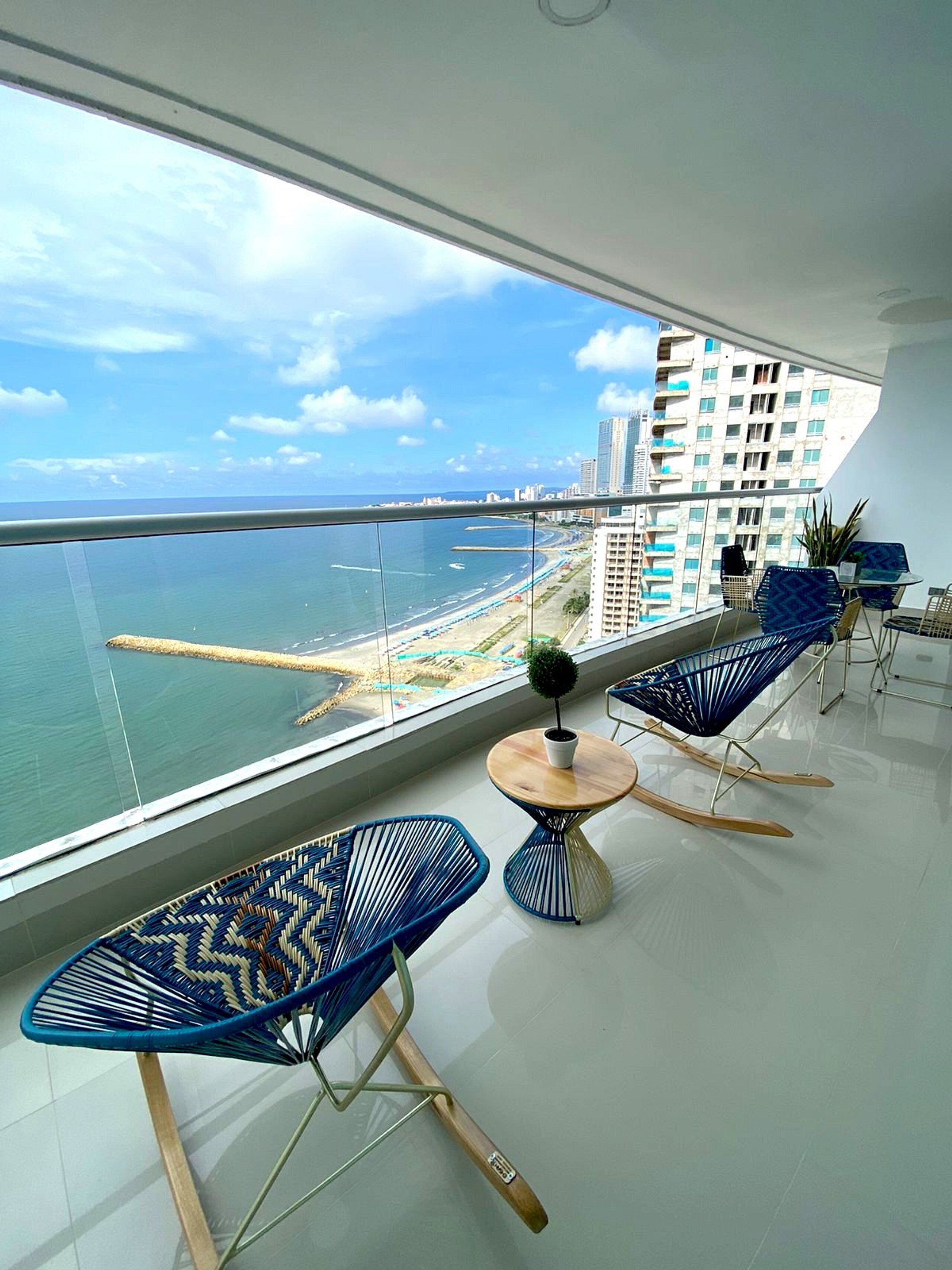 Un balcon avec vue sur la mer et sur le paysage urbain, aménagé avec des chaises bleues et une petite table.