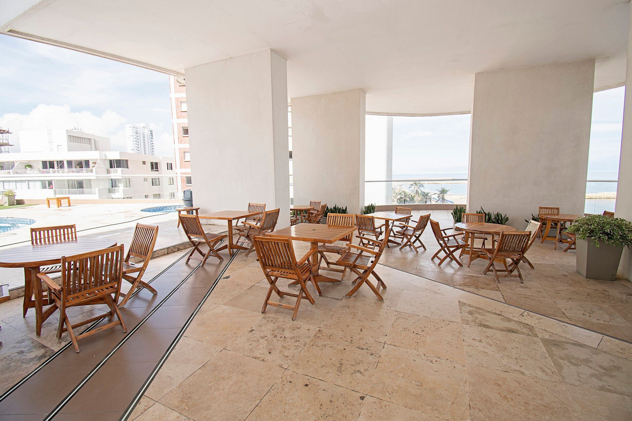 A spacious outdoor seating area with multiple wooden tables and chairs, large windows offering a view of the cityscape and beach, and a tiled floor.