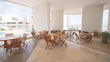 A spacious outdoor seating area with multiple wooden tables and chairs, large windows offering a view of the cityscape and beach, and a tiled floor.