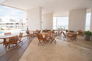 A spacious outdoor seating area with multiple wooden tables and chairs, large windows offering a view of the cityscape and beach, and a tiled floor. - U-GO Edificio Palmetto Eliptic Cartagena (Cartagena)