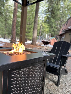 A fire feature with a glass of red wine on a metal table, surrounded by outdoor chairs. - Cozy Pines Cabin (Ronald)