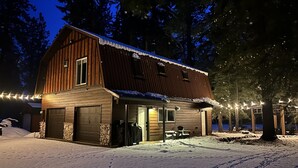 A wooden barn-style building with a garage, surrounded by snow-covered ground and trees. - Cozy Pines Cabin (Ronald)