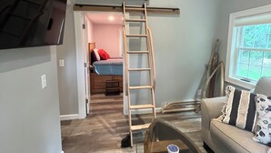 Un dormitorio tipo loft con cama en entrepiso, escalera de madera, una mesa de centro de vidrio y un sofá con cojines estampados.