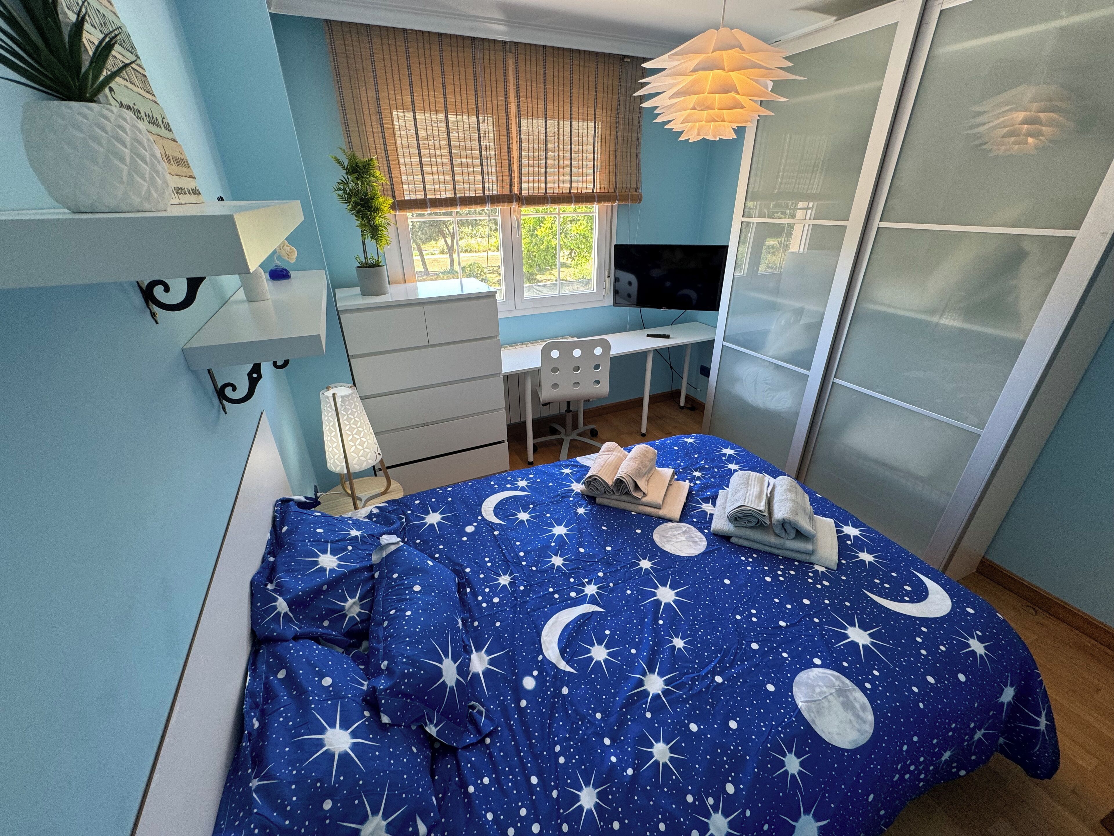 Une chambre à coucher avec un lit recouvert d’une couette à motifs de ciel étoilé, un bureau avec un ordinateur, une télévision et une fenêtre avec des stores.