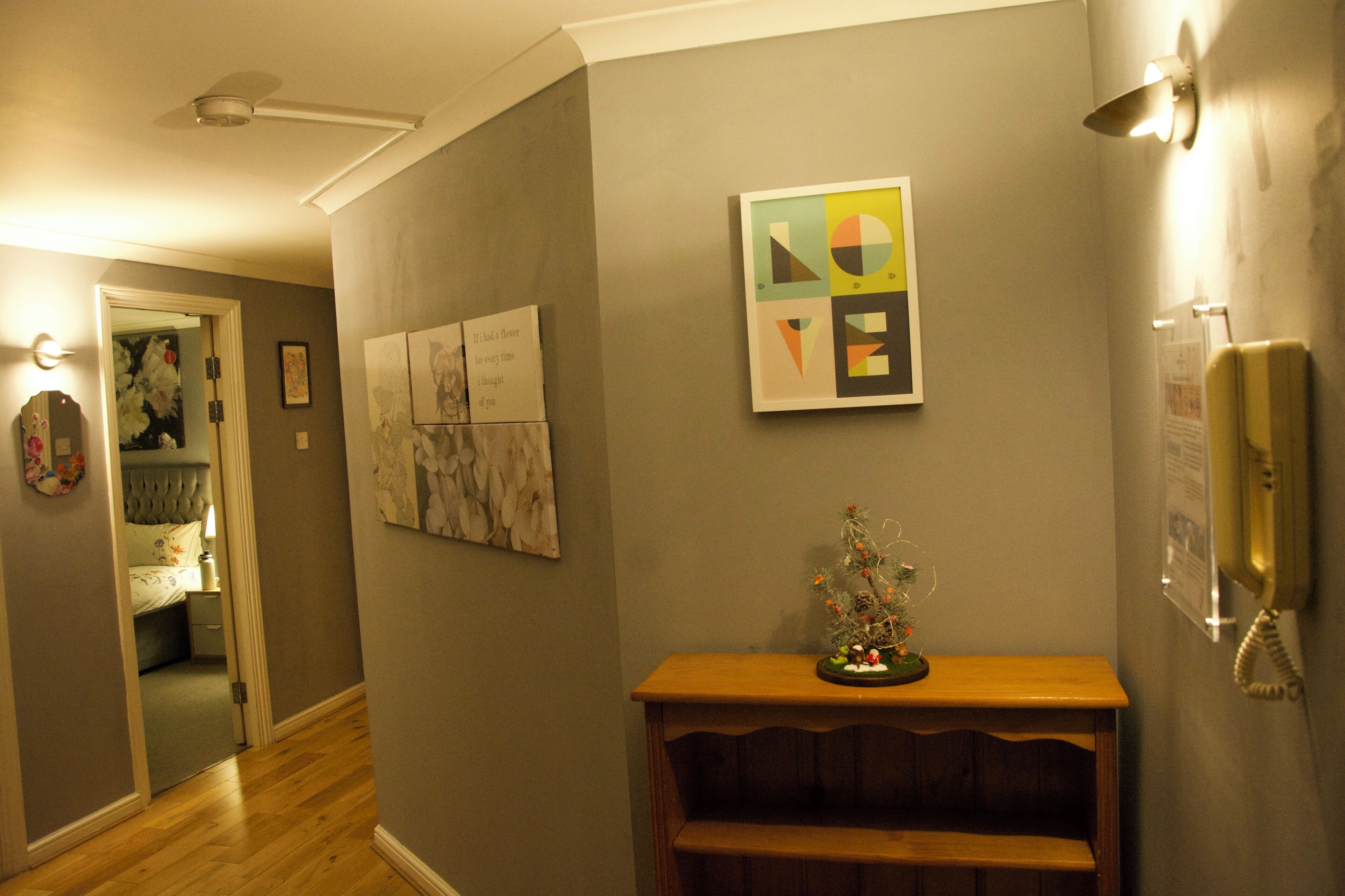 A hallway with a wooden floor, a small wooden table with a decorative item, and a wall with framed artwork.