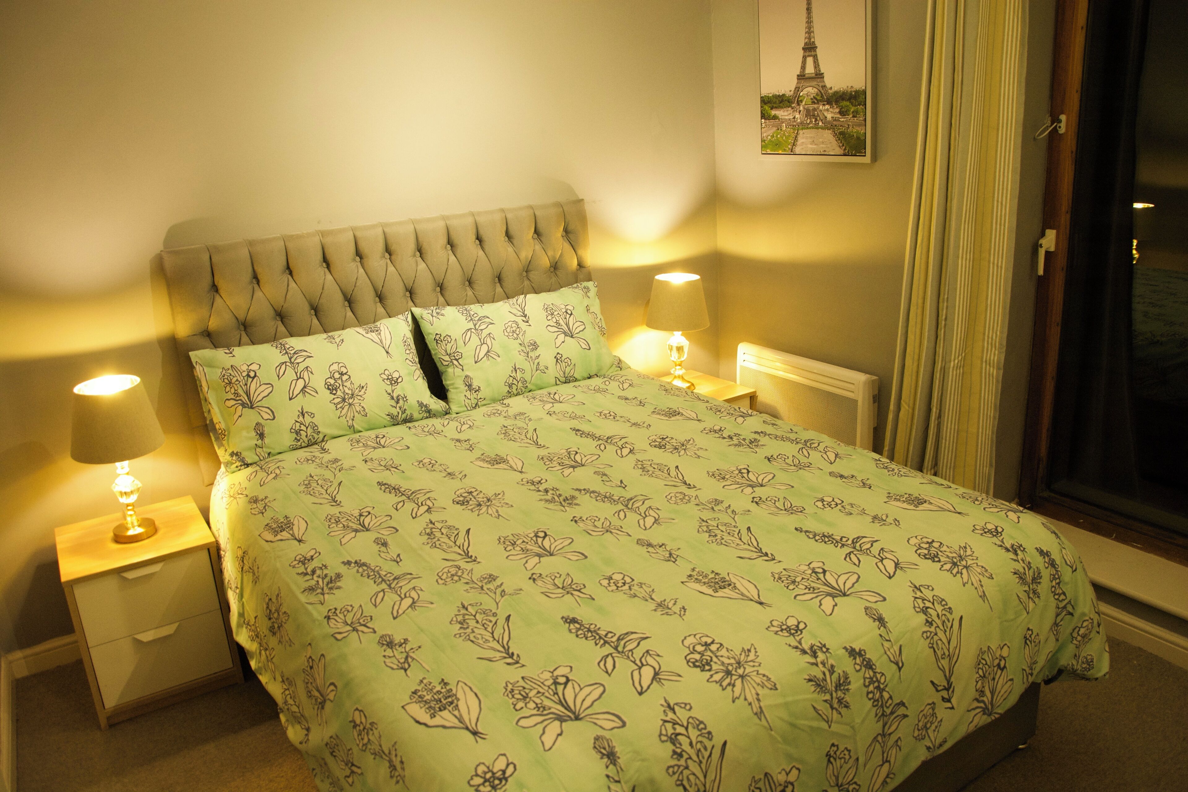 Una cama bien hecha con ropa de cama floral, dos lámparas de noche y vistas a la Torre Eiffel a través de una ventana.
