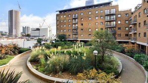 A modern apartment building with multiple balconies, a landscaped garden with various plants, and a view of the cityscape in the background. - Lux 2 Bed in Canary Wharf Close to Excel & O2 (London)