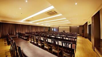 A conference room with rows of chairs and a long table, equipped with ceiling lights and a large screen.
