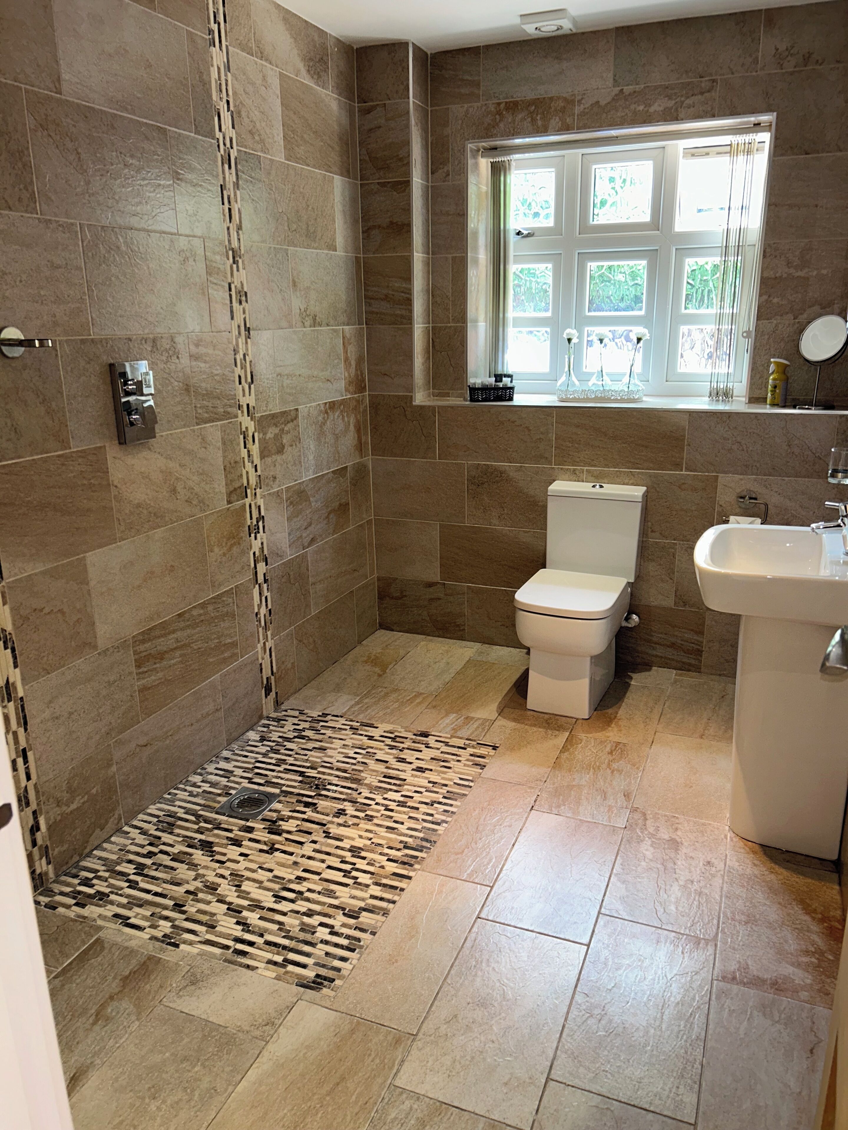 A bathroom with a shower, toilet, and sink, tiled with beige tiles and a patterned rug.