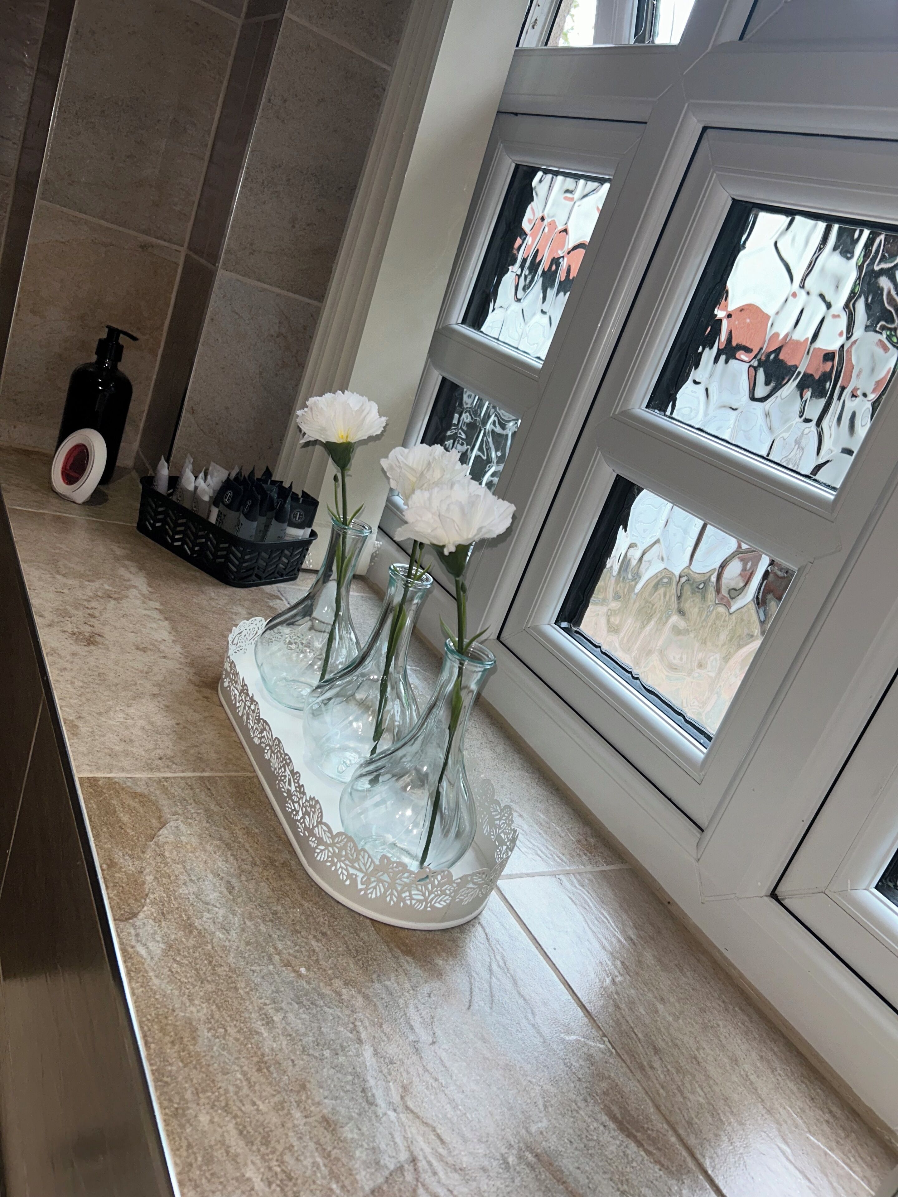 A bathroom with a tiled floor, a black soap dispenser, a basket of toiletries, and three white flowers in glass vases on a decorative tray.