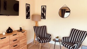 A living room with a wooden cabinet, two striped armchairs, a small table with a plant, a floor lamp, and a TV mounted on the wall. - Claverley Hideaway Apartment Bridgnorth (Wolverhampton)