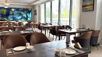 Un comedor con mesas dispuestas para el servicio, ventanas amplias y un cuadro en la pared.