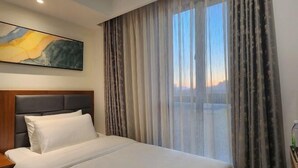 Ein Hotelzimmer mit Bett, Kopfteil, einem Gemälde an der Wand und einem Fenster mit Vorhängen.