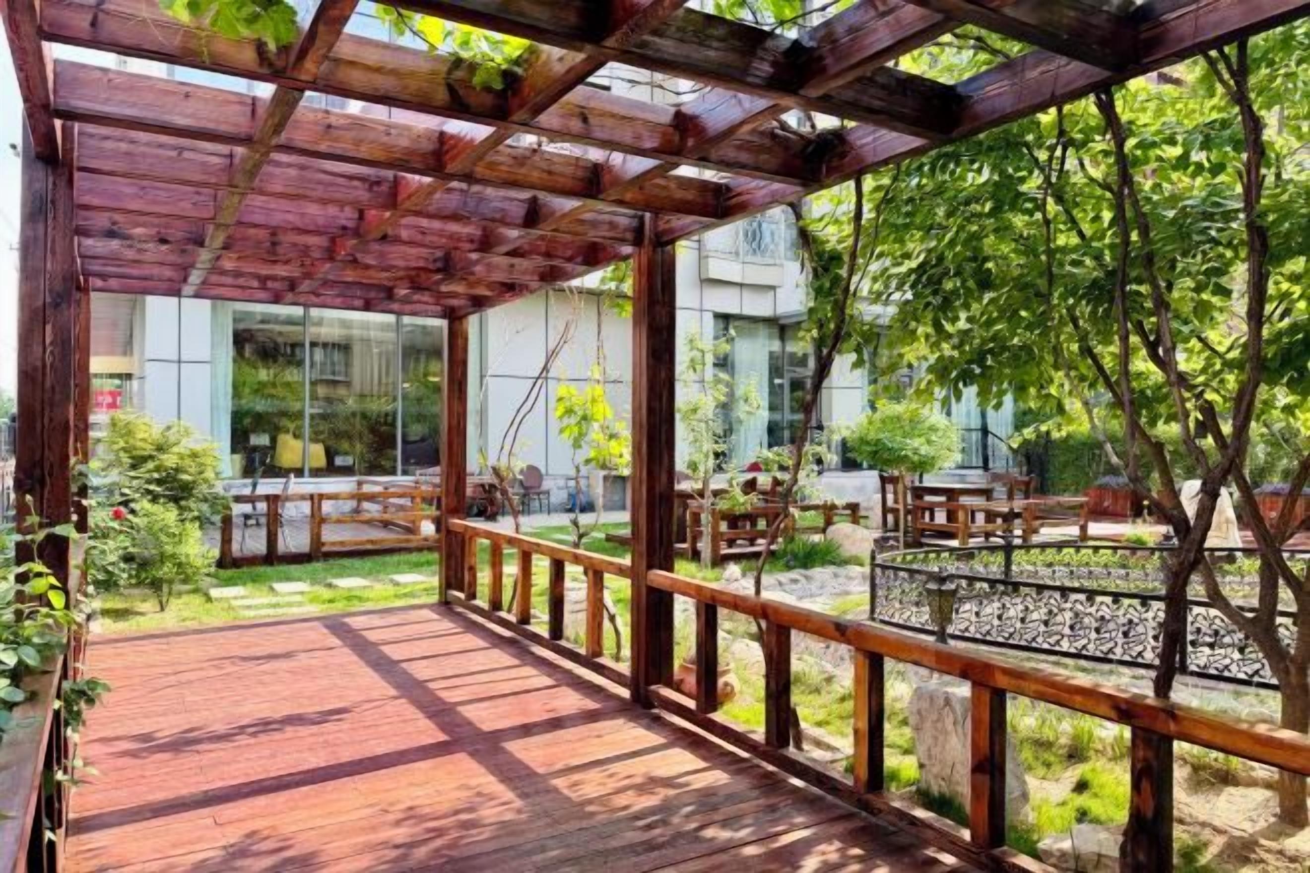 A wooden deck with a railing, surrounded by greenery and a building in the background.