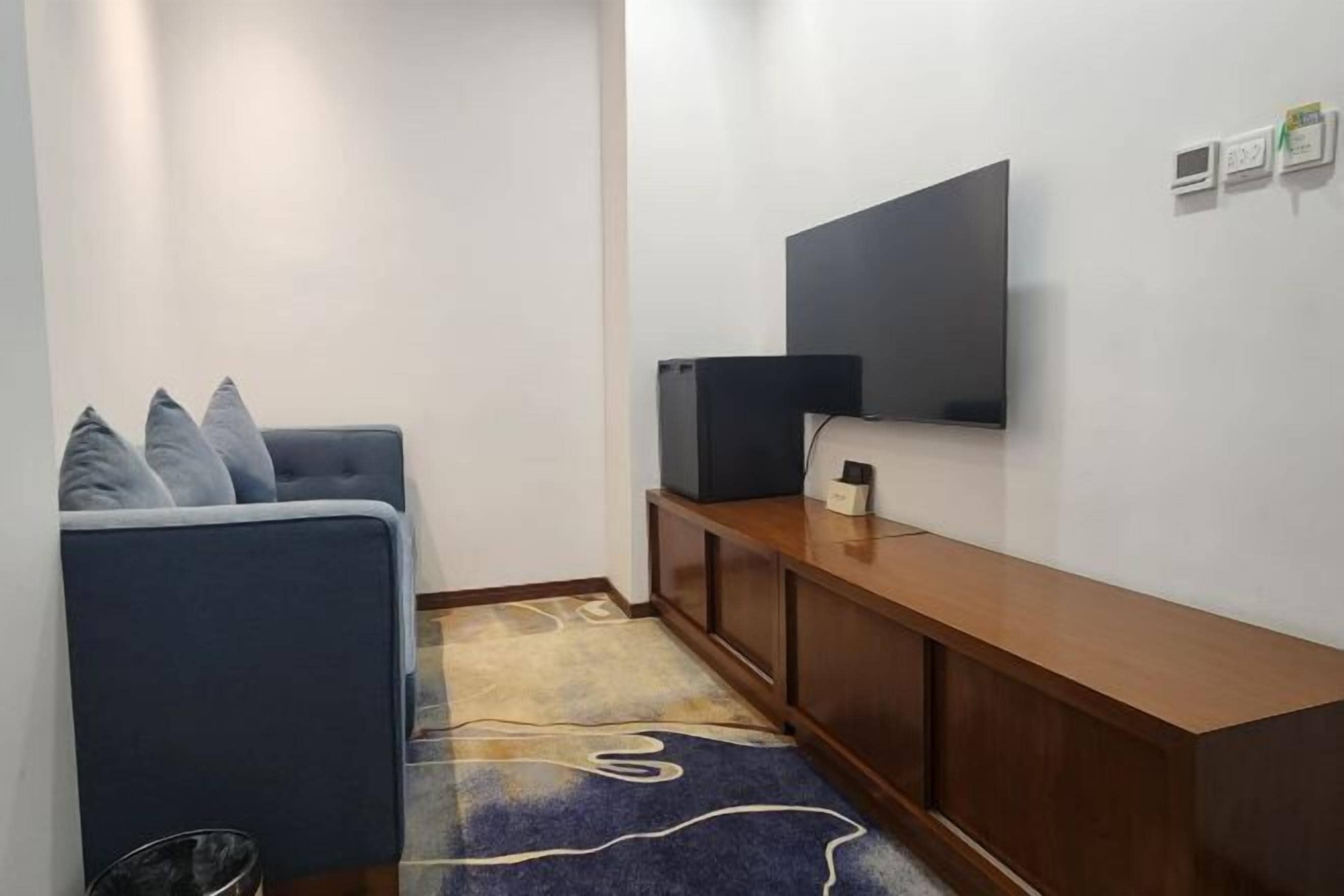 Una sala pequeña con un sofá, un televisor montado en la pared, una consola de madera y un tapete estampado.