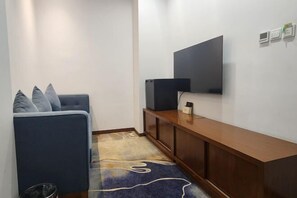 Uma pequena sala de estar com um sofá, uma televisão fixada na parede, uma mesinha de console de madeira e um tapete estampado.