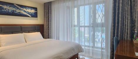 A hotel room with a large bed, a wooden headboard, a dining table, and a view through sheer curtains.