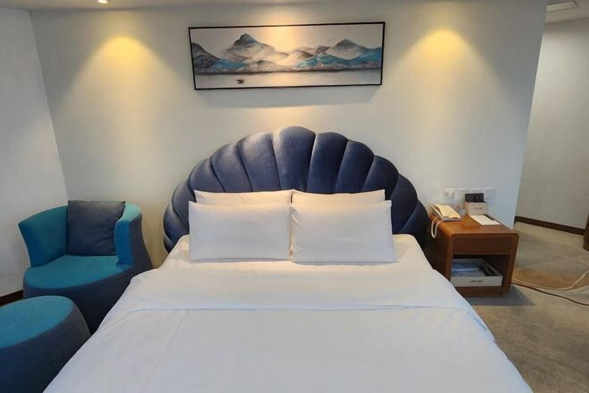Uma cama com cabeceira original, uma mesa de cabeceira com abajur, uma cadeira e um quadro na parede.