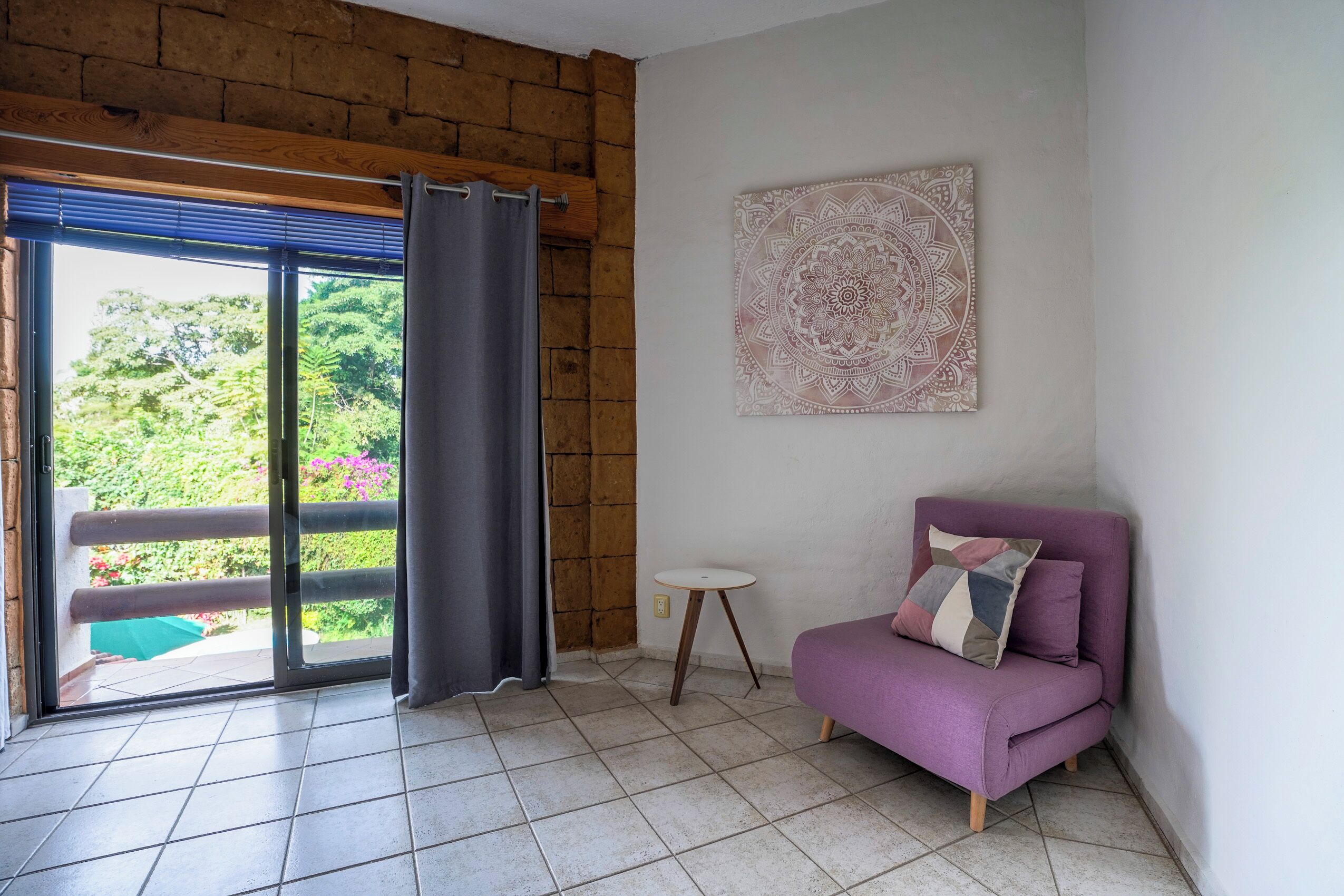 Een kamer met een paarse bank, een kleine ronde tafel, een groot raam met gordijnen en een decoratief object aan de muur.