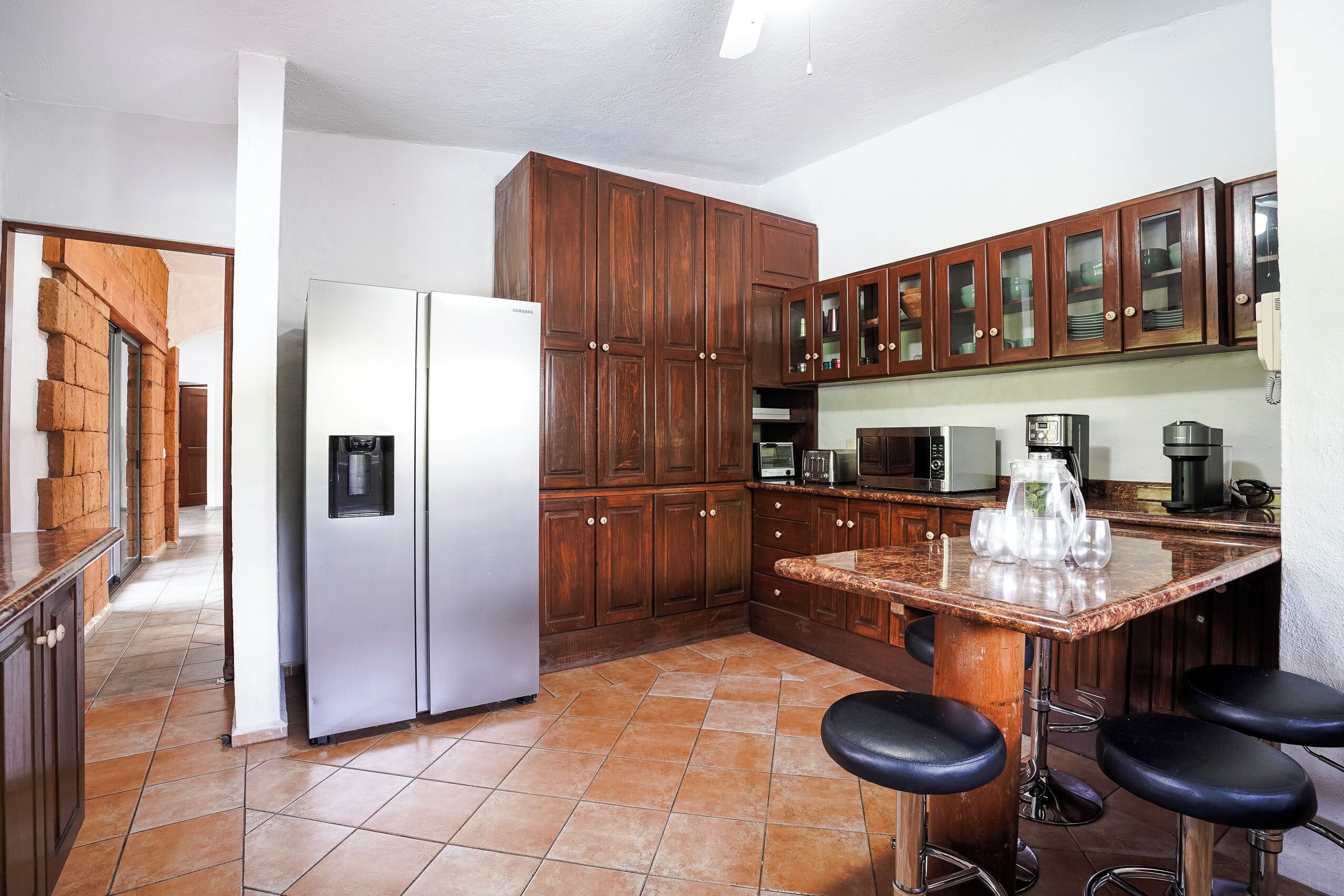 A kitchen with wooden cabinets, a refrigerator, a microwave, and a bar area with stools.