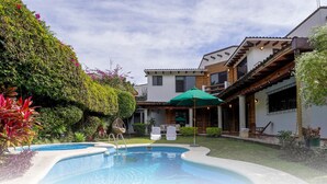 Un área de piscina con una zona de estar techada y un edificio de techo con tejas.
