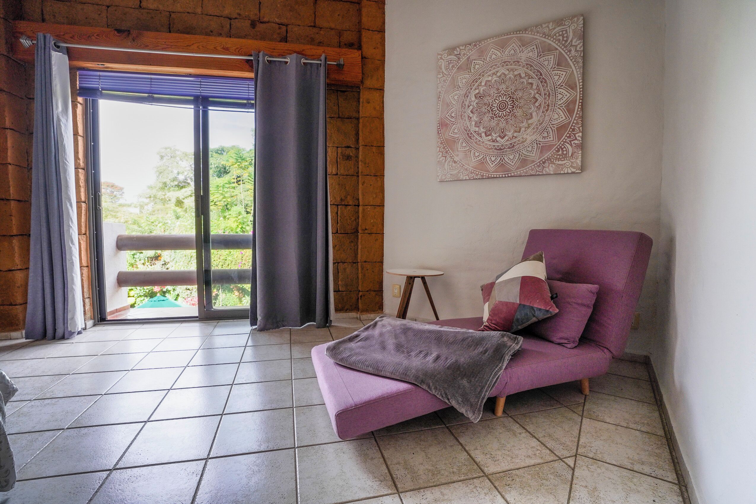 A room with a purple sofa, a blanket, a small table, and a wall with a mandala design.