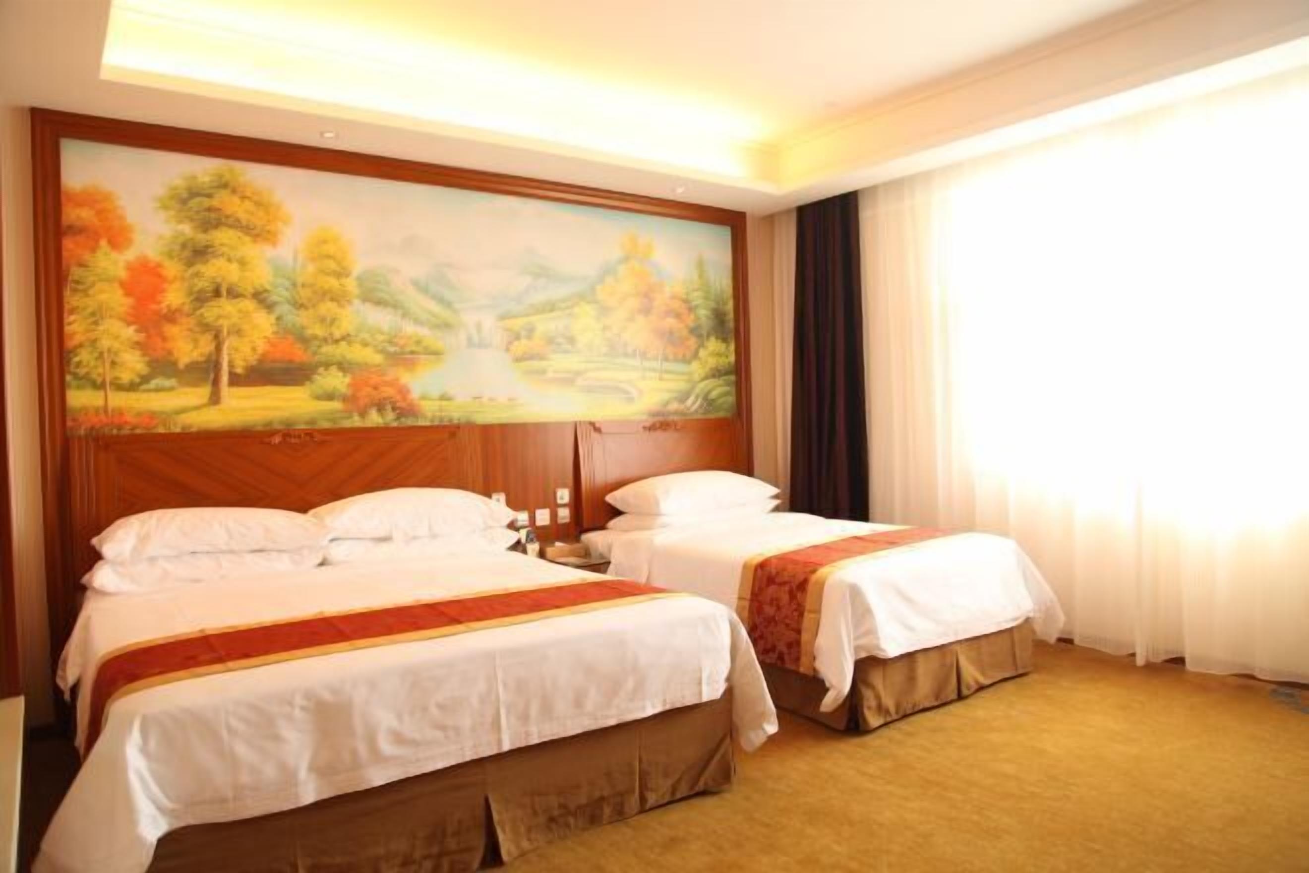 A hotel room with two beds, a painting of a landscape, and a large window with curtains.