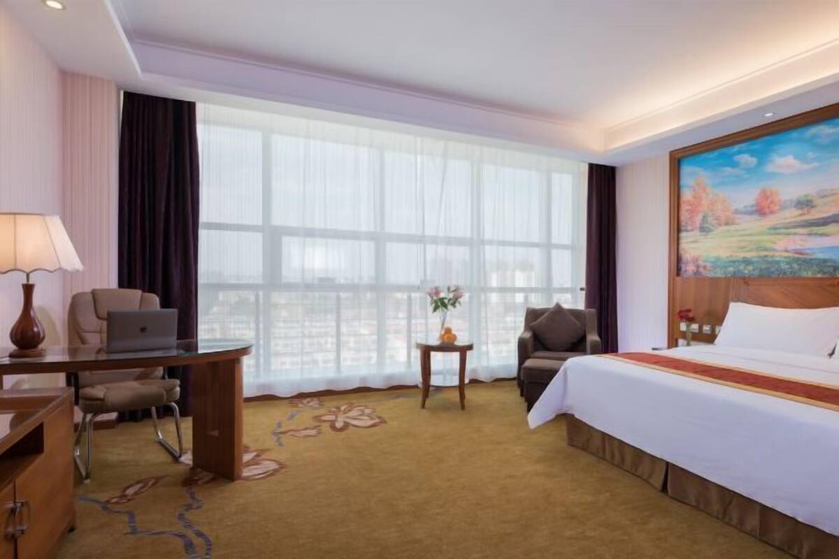 Hotellihuone, jossa on suuri ikkuna, työpöytä kannettavalla tietokoneella, sänky ja taulu seinällä.