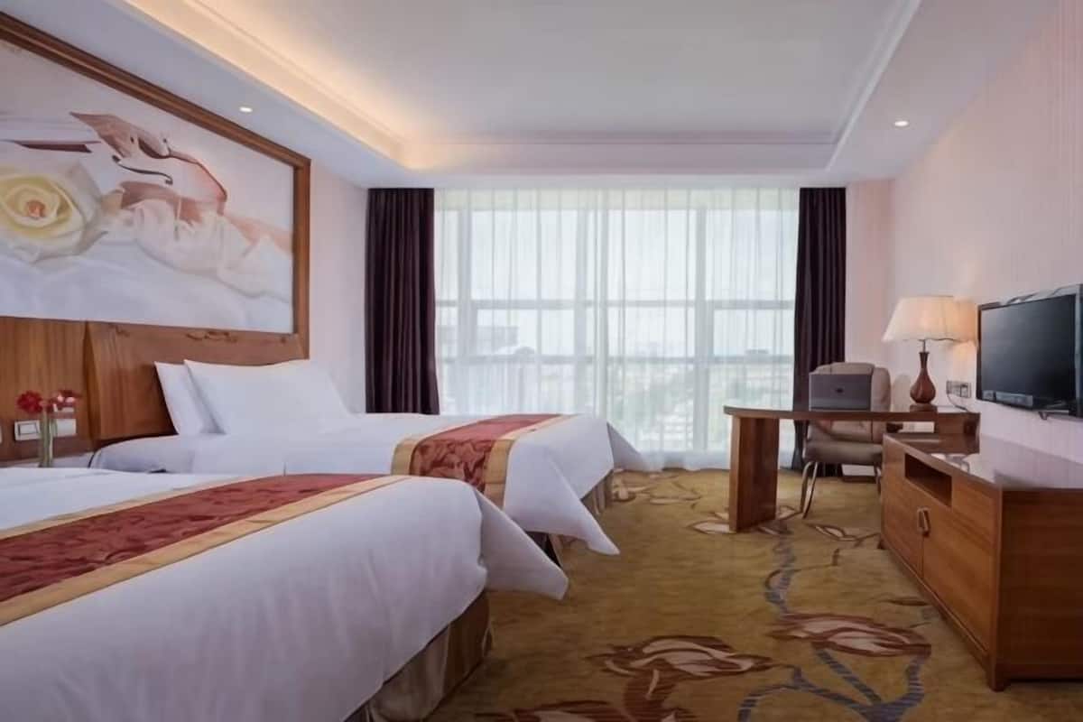 A hotel room with two beds, a television, a desk with a lamp, and a large window.