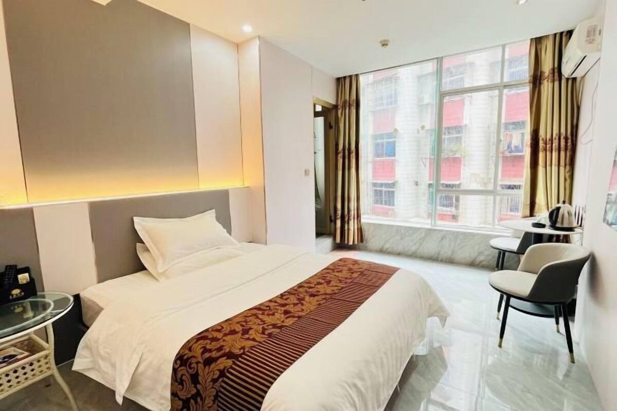 A hotel room with a bed, a desk, a chair, a window with curtains, and a view of buildings outside.