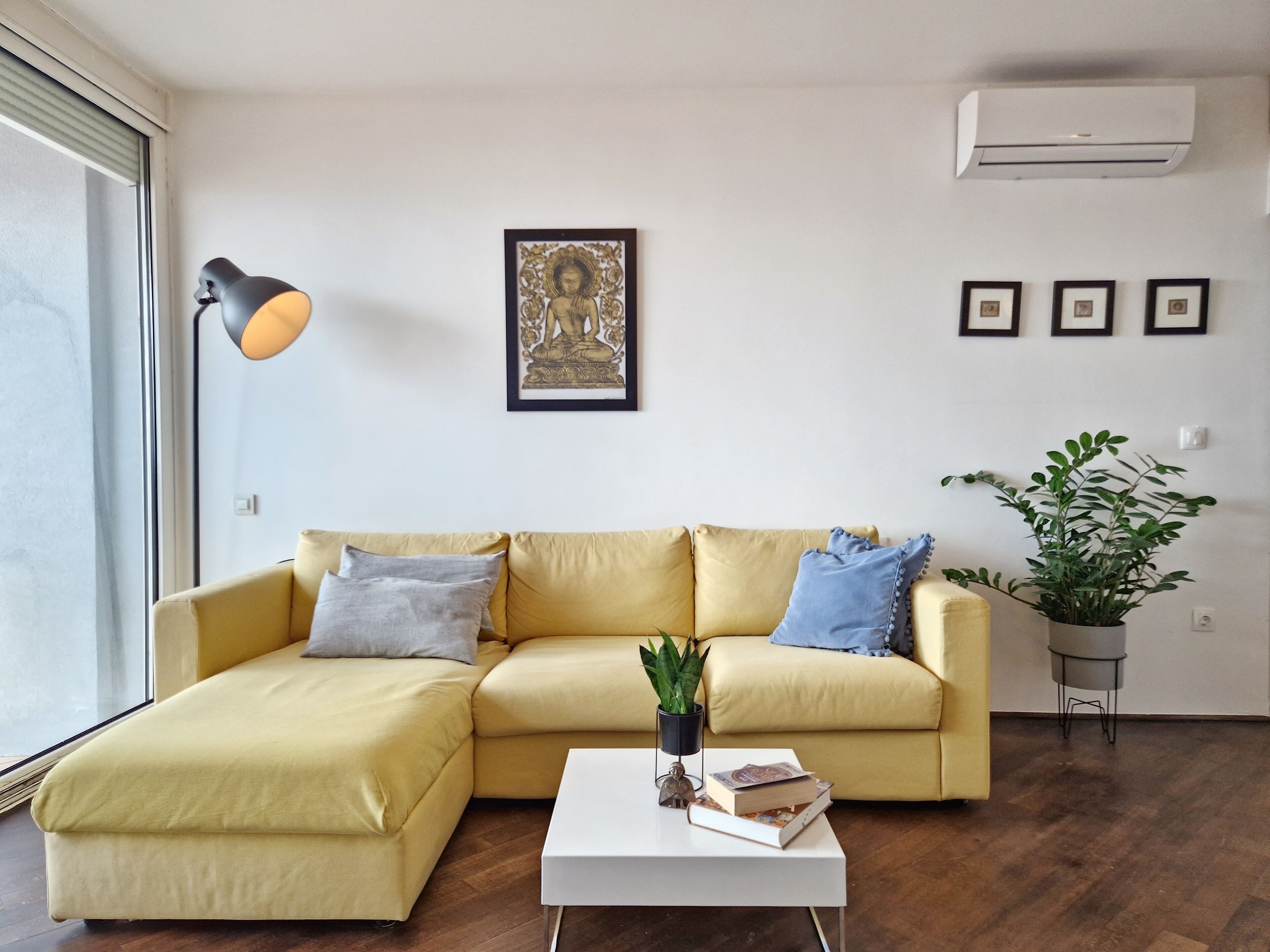 Un salón con un sofá amarillo, una mesa de centro blanca con libros, un cuadro enmarcado en la pared y una planta en maceta.