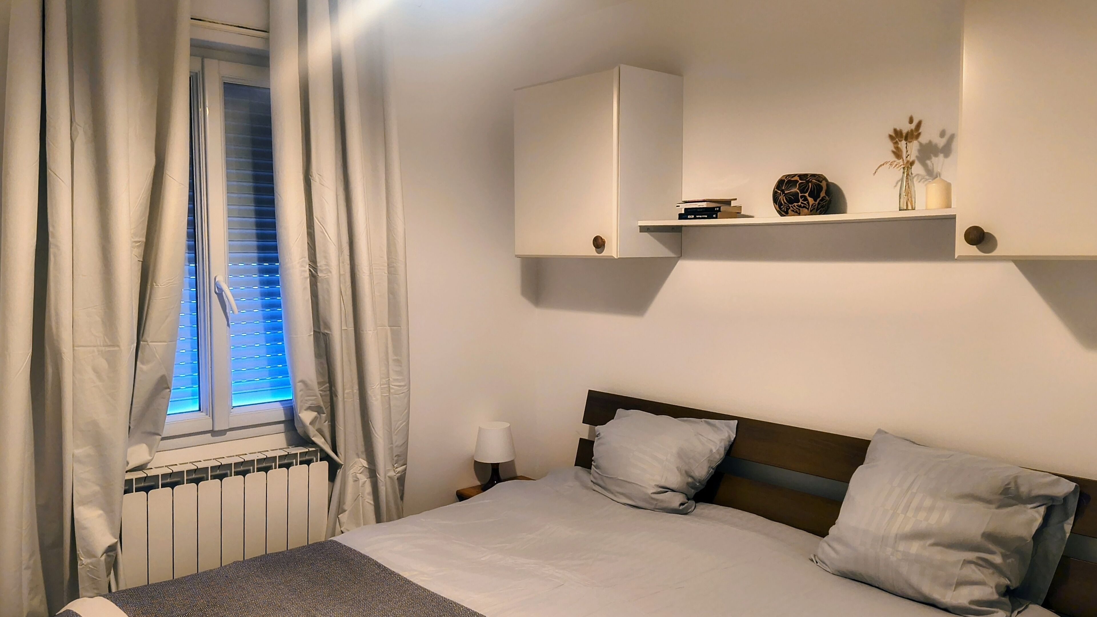 Ein Schlafzimmer mit einem Bett, einem Nachttisch, einem Fenster mit Vorhängen, einem Heizkörper und einem Regal mit Dekoration.